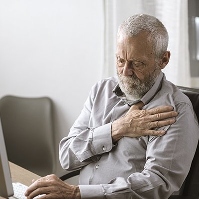 Man sitting at a laptop clutching at this chest with one hand 